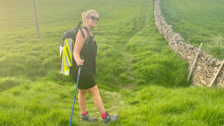 A person standing on a grassy, green country path in the UK, wearing a hiking boots, walking poles a day pack and sunglasses.