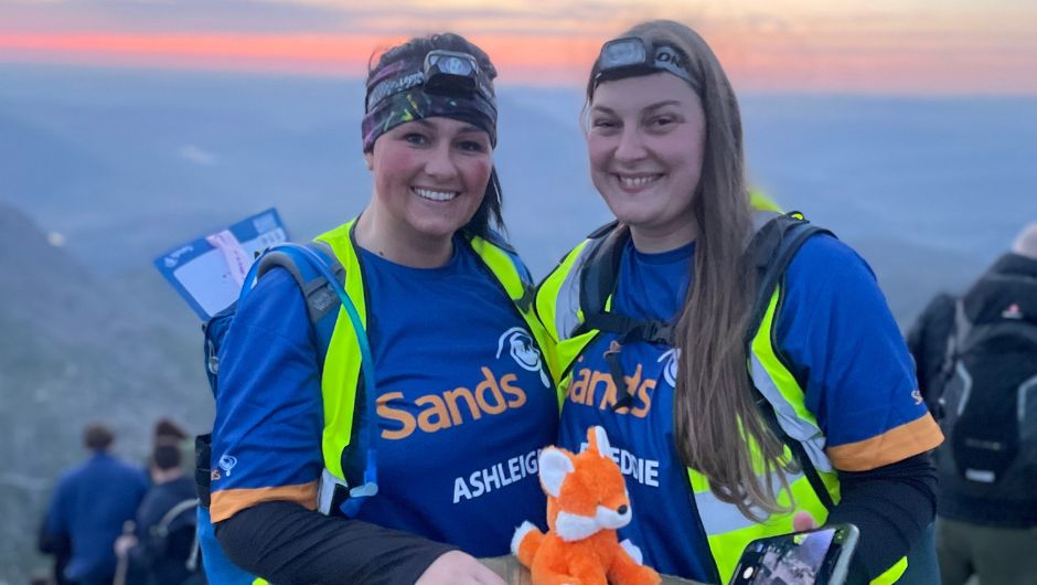 Two smiling women wearing headlamps and high-visibility vests pose at a mountain viewpoint during sunrise, raising money for Sands charity and holding a small orange plush toy.