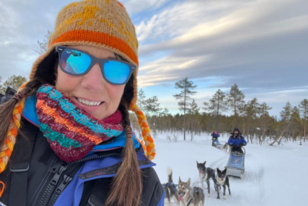Smiling woman wearing winter clothing and sunglasses takes a selfie while dog sledding through a snowy forest, with huskies and sled visible behind her.