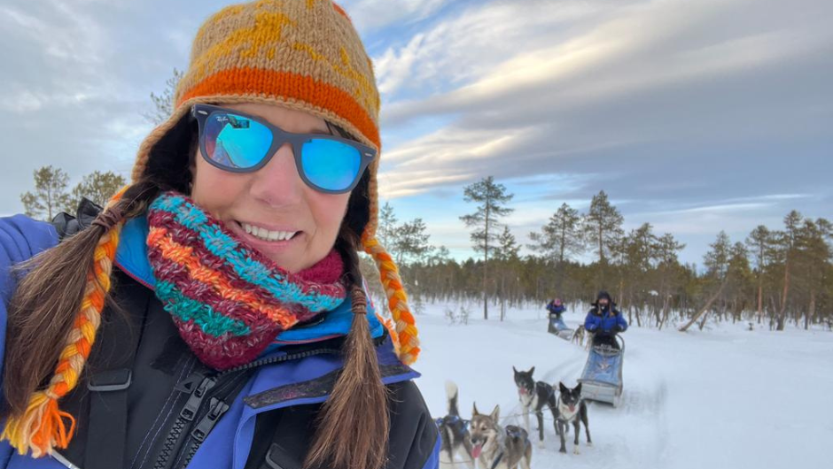 Smiling woman in winter clothing taking a selfie while dog sledding through a snow-covered forest in Swedish Lapland, with huskies pulling sleds behind her.