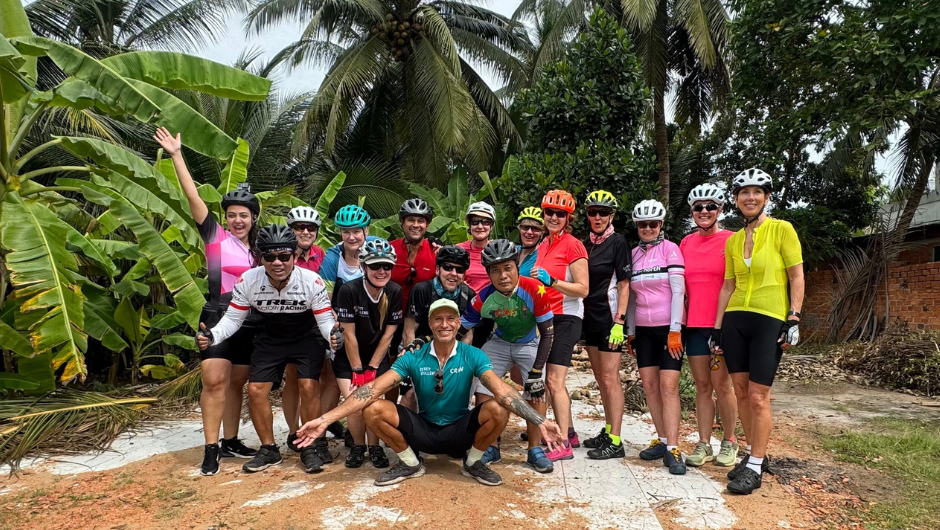 Group of cyclists on the Saigon to Angkor Wat bike ride, posing on a rural road surrounded by palm trees in South East Asia.