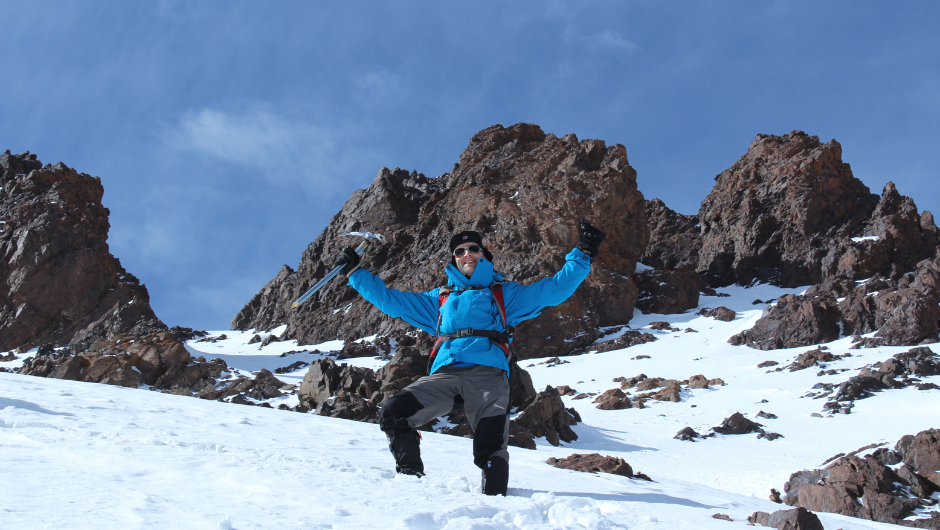Trekker celebrating on a snowy slope during a winter ascent of Mount Toubkal in Morocco’s High Atlas Mountains, surrounded by rugged rocky peaks under a clear blue sky.