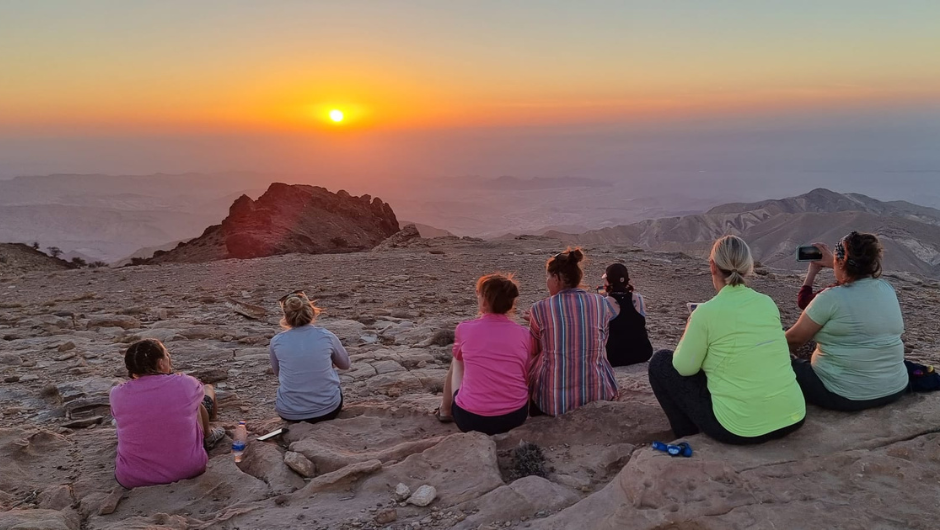 a group of people sat on a rocky ledge in the jordan desert, looking out at a glowing orange sunset.