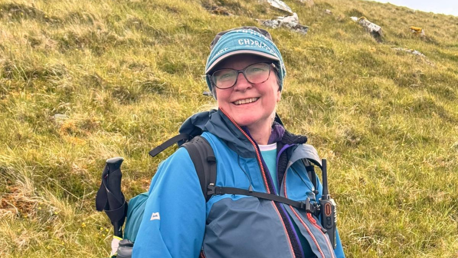 A challenge leader in front of a grassy hill, smiling, wearing a rain coat, day pack and a charity challenge neck buff as a headband