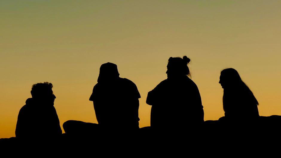 Four silhouetted people sitting beneath an orange sunset sky, talking together.
