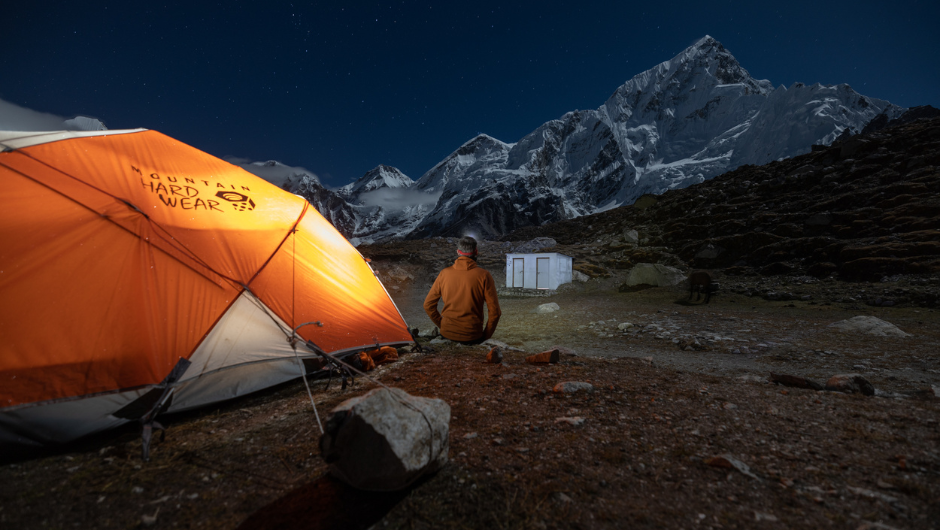 A person sat outside an orange tent at nighttime, looking up at a Himalayan mountain range and a starry night sky.