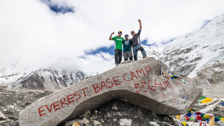 Three people celebrating on top of a large stone that has Everest Base Camp 5364m written on it. Snowy Himalayan peaks are in the background.