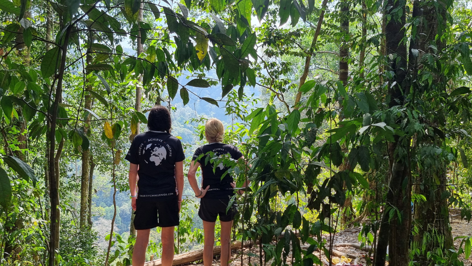 Two trekkers wearing Charity Challenge t-shirts, looking out at a lush green rainforest view, surrounded by the Sumatran rainforest canopy.