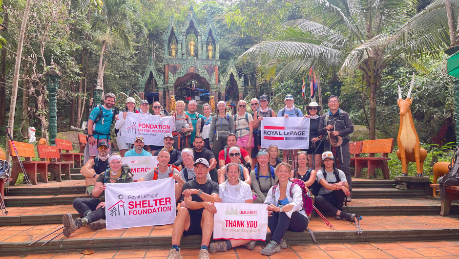 A large group of hikers in casual of an ornate gateway and lush greenery, holding banners that say "Royal Lepage Shelter Foundation."