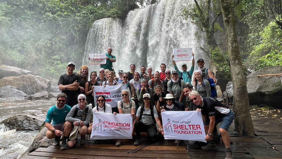 A large group of hikers pose for a photo in front of a lush waterfall, holding banners that read "Thank You" "Merci" and "Royal LePage Shelter Foundation."