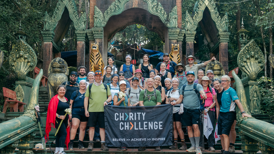 A large group of smiling people posing for a photo in front of a temple entrance featuring tiger statues and serpent motifs, holding a banner that reads "CHARITY CHALLENGE BELIEVE >> ACHIEVE >> INSPIRE"