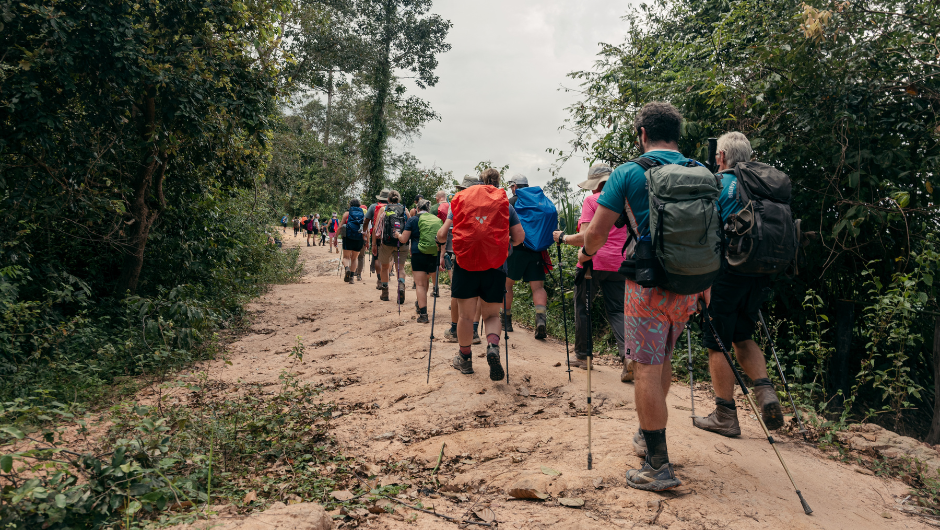 A group of hikers with backpacks walk along a dirt path surrounded by lush green vegetation under a cloudy sky.