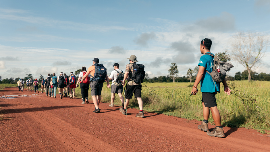A group of hikers with backpacks trekking along a dirt path in Cambodia, next to some lush green fields.