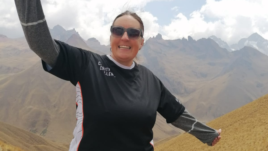 A woman wearing a Charity Challenge T-Shirt, smiling for the camera against a backdrop of the Andes Mountains in Peru.