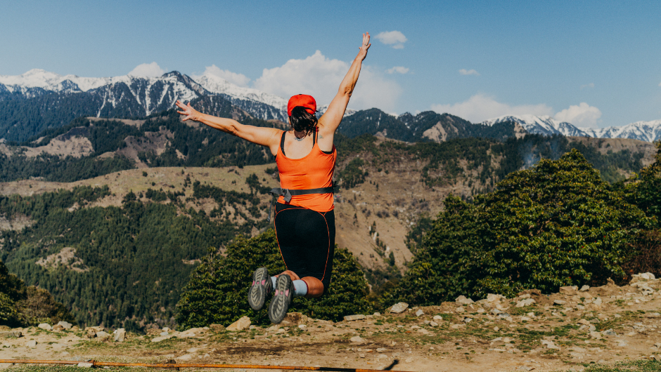 A woman dressed in trekking gear jumping up in celebration, looking out at a view of the Indian Himalayan Mountains.