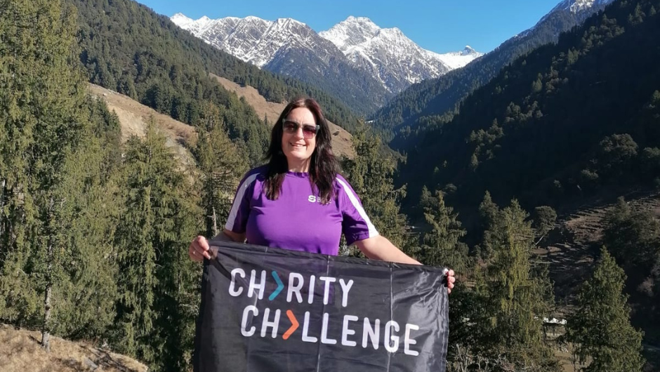 Sharon Myers, a Charity Challenge participant, holding up a Charity Challenge flag against a backdrop of snow-capped Himalayan mountains and forested hills in India.