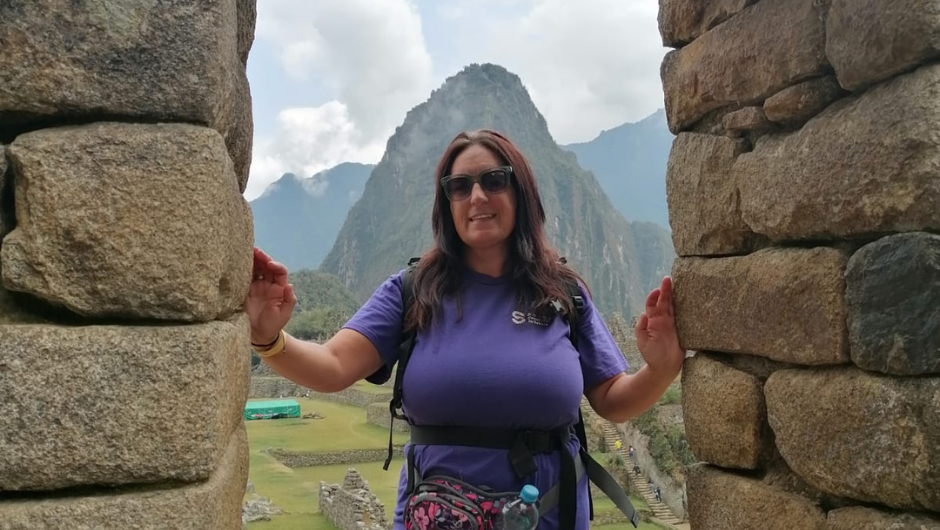 Sharon Myers Challenger Stories: Sharon stood at the site of Machu Picchu, in an archway of an Incan stone building, with a mountain peak behind her.