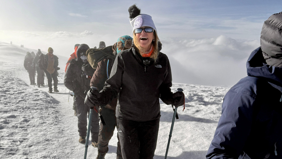 A person smiling at the camera as they trek up a snowy mountain path with a group of other trekkers.