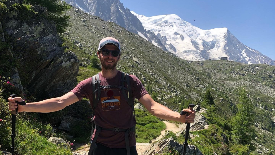A person smiling on a sunny mountain path, holding two walking poles beside them. There are snow-capped mountain peaks in the background.