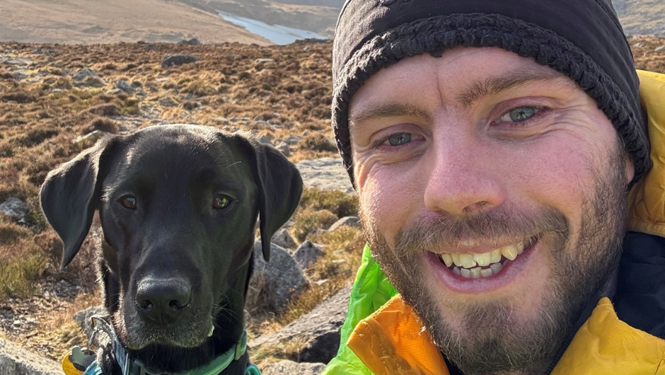 A person smiling in a selfie with his dog, surrounded by marshy land.