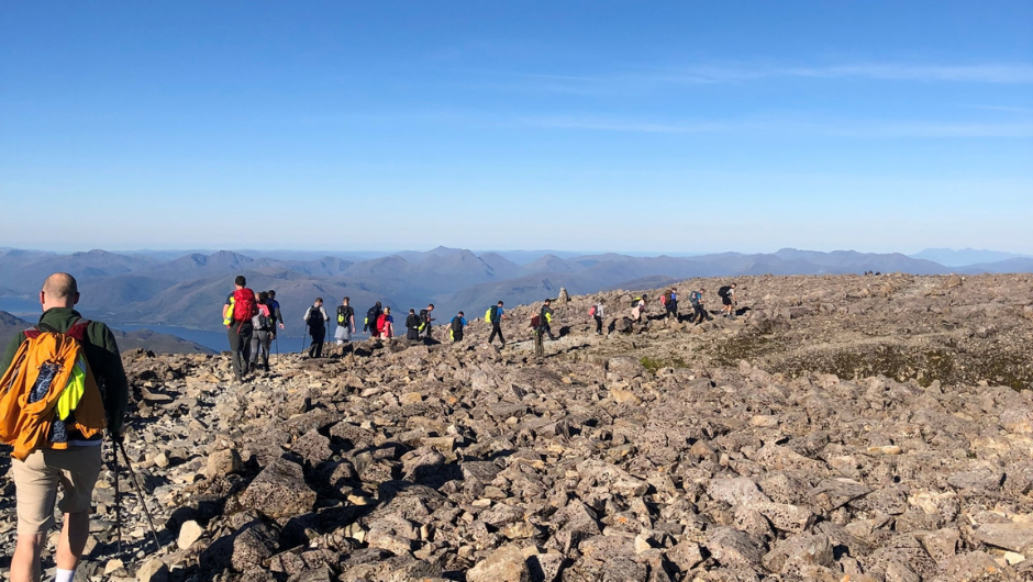 A line of trekkers walking across a flat rocky section of a mountain. A lake and mountain peaks are in the far distance.
