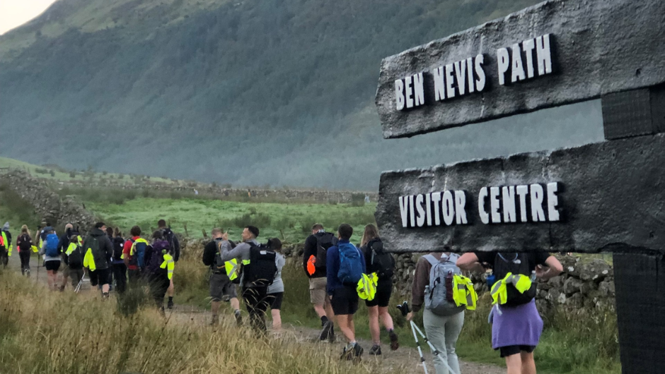 A line of trekkers walking along the Ben Nevis Path, with a wooden Ben Nevis Path sign post in the foreground.
