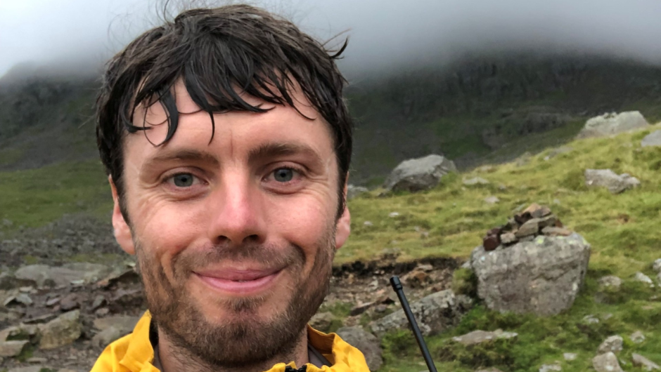 A person smiling surrounded by a grassy and rocky mountain path.