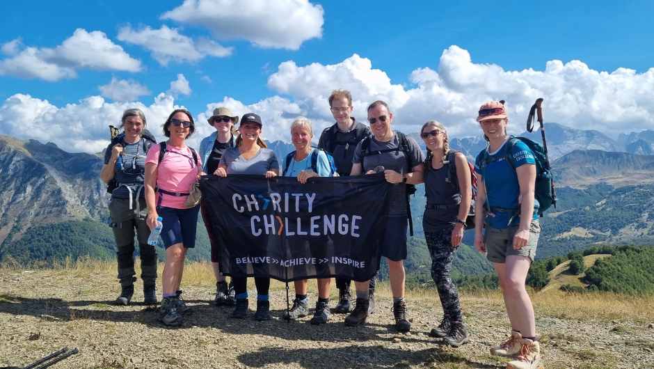 A group of trekkers stood in front of a mountain range and blue skies holding up a charity challenge flag.