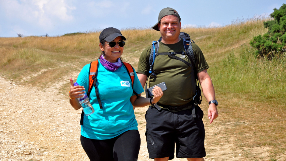 Two people dressed in trekking clothes, wearing caps and backpacks, walking along a country path and smiling for the camera.