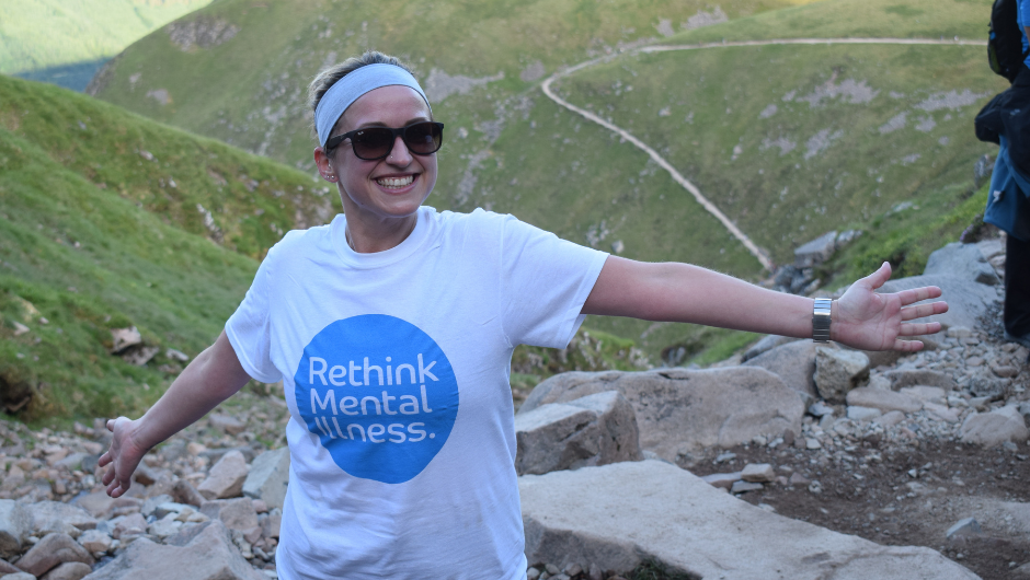 A person wearing a 'Rethink Mental Illness' t-shirt and smiling at camera, as she walks up a UK mountain path.