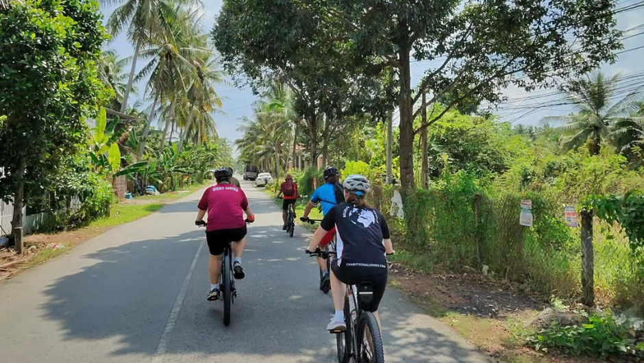 a group of people cycling along a pathed road in south east Asian, lined by green trees.