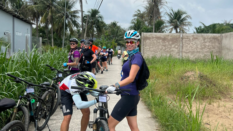A smiling cyclist holding her bike upright as another cyclist helps adjust the bike seat.