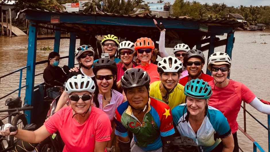 A group of cyclists wearing helmets and smiling together as they pass the Mekong river on a small ferry.