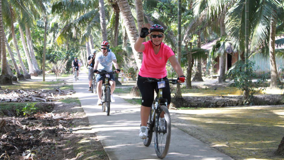A single line of cyclists riding on a narrow pathed path, surrounded by palm trees.