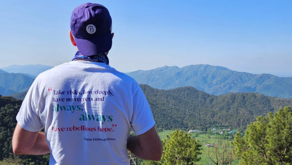 A person wearing a Bowelbabe Fund t-shirt and cap, facing out towards a green mountain range.