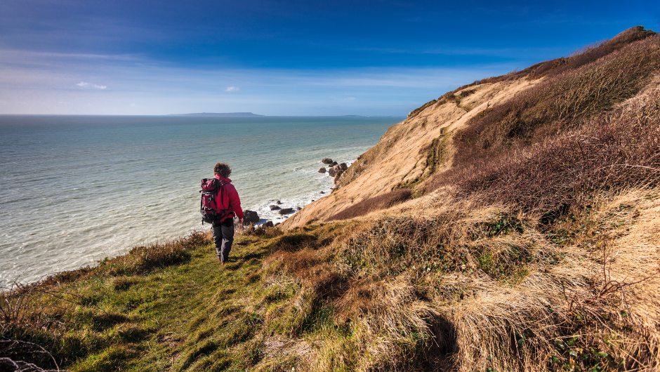 A person wearing a rain jacket and backpack, walking along a coastal path next to the sea.