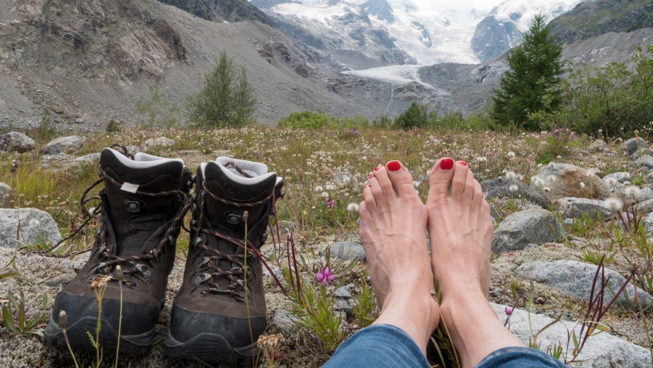 a pair of bare feet, with a pair of open walking boots next to them, perched on a rock, with a mountain view in the background.