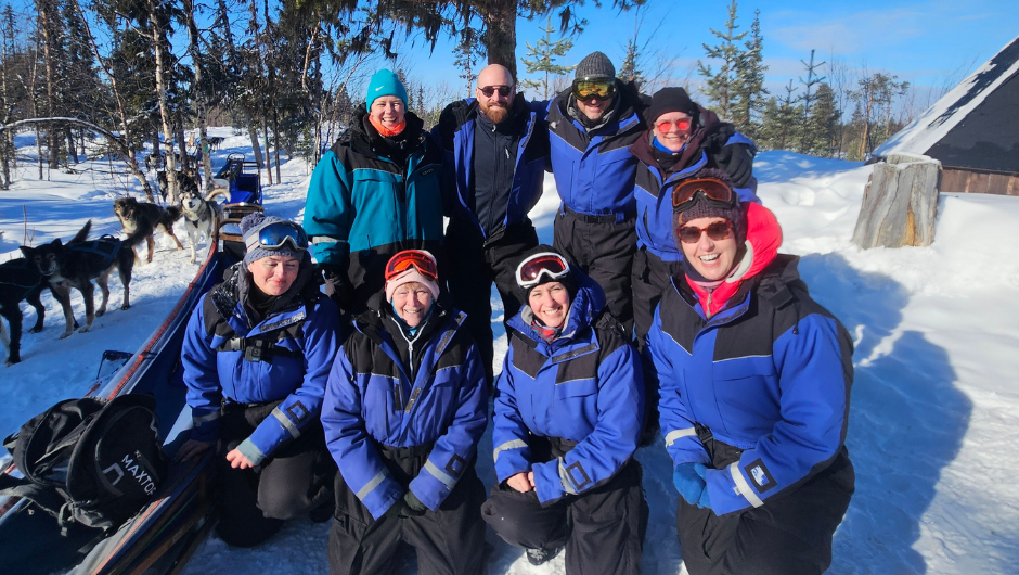 A group of Dog Sledding Challenge charity challenge participants smiling for a group photo, dressed in their snow suits and goggles, with some huskies in the background.