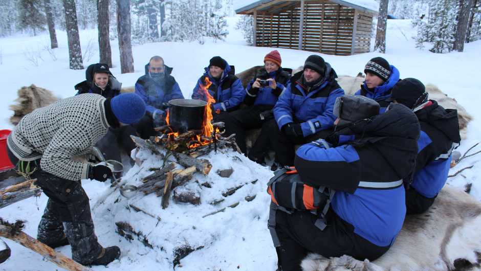 A group of people wearing snowsuits, sitting outside in the snow, surrounding a fire with large pot on top of it.