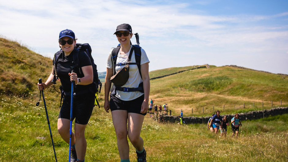 two trekkers smiling for the camera, with a section of hadrian's wall behind them