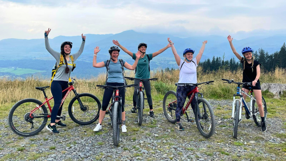 A group of people standing stationary with their mountain bikes, raising their arms in celebration, during their lake district triple challenge