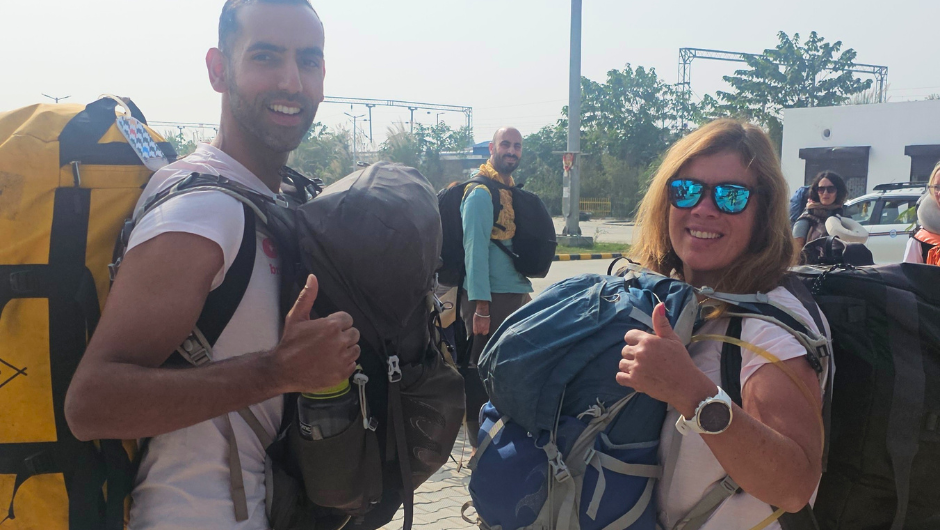 Two people smiling with their thumbs up for the camera, wearing a large bag on their back and a daypack on their front.