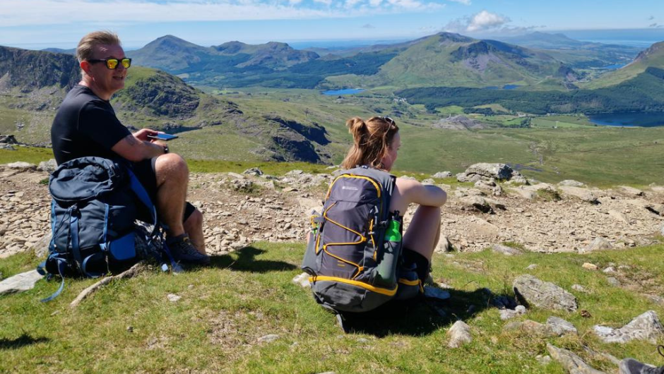 Two people sitting to rest during their trek, over looking green hills below.