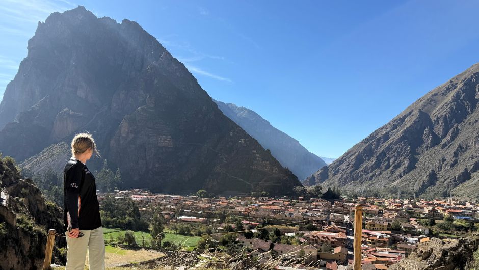 A person standing in front of a tall rocky mountain, with a valley and village buildings in the foreground.