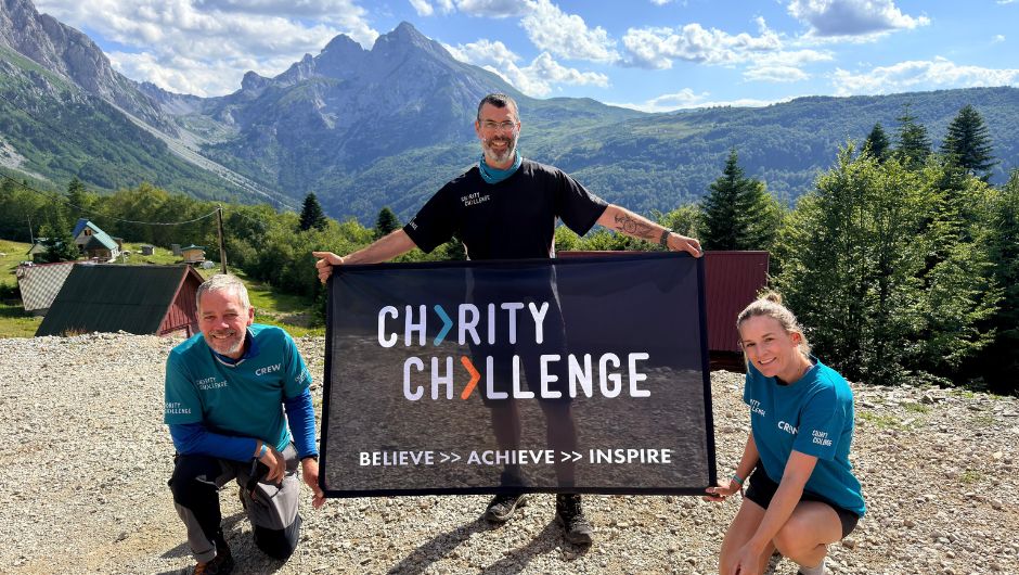 Three people holding up a Charity Challenge logo flag, in front of a mountainous background.